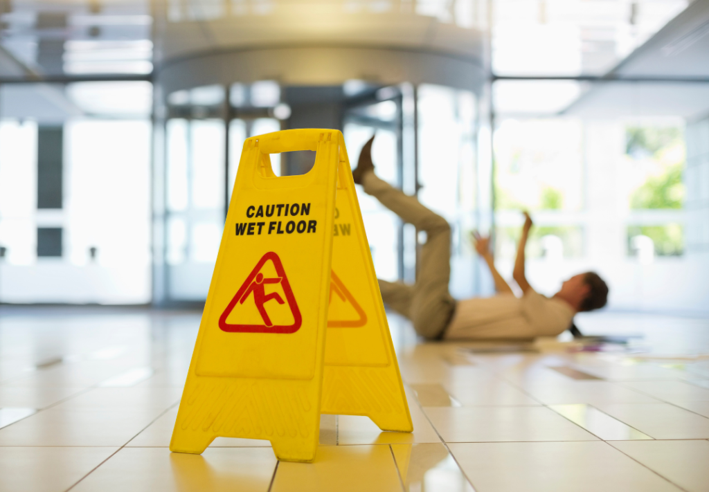 A caution "wet floor" sign in the foreground with a blurred image of a man slipping and falling in the background inside a modern store in California, emphasizing the Steps to Take If You Slip and Fall in a California Store to protect your legal rights.
